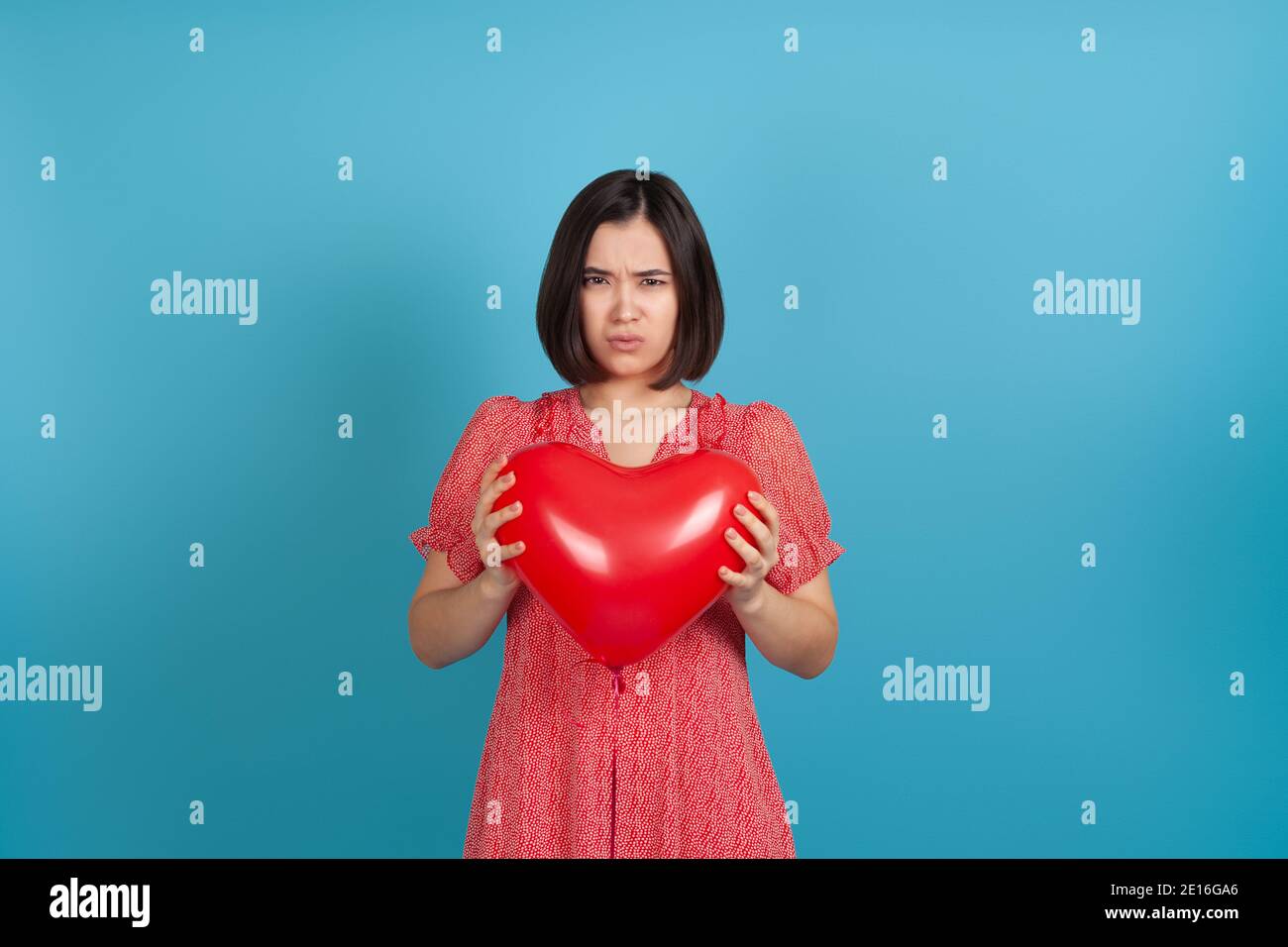 mock up offended by a bad gift, a young Asian woman holds a red heart-shaped balloon in her hands on Valentine's day, isolated on a blue background Stock Photo