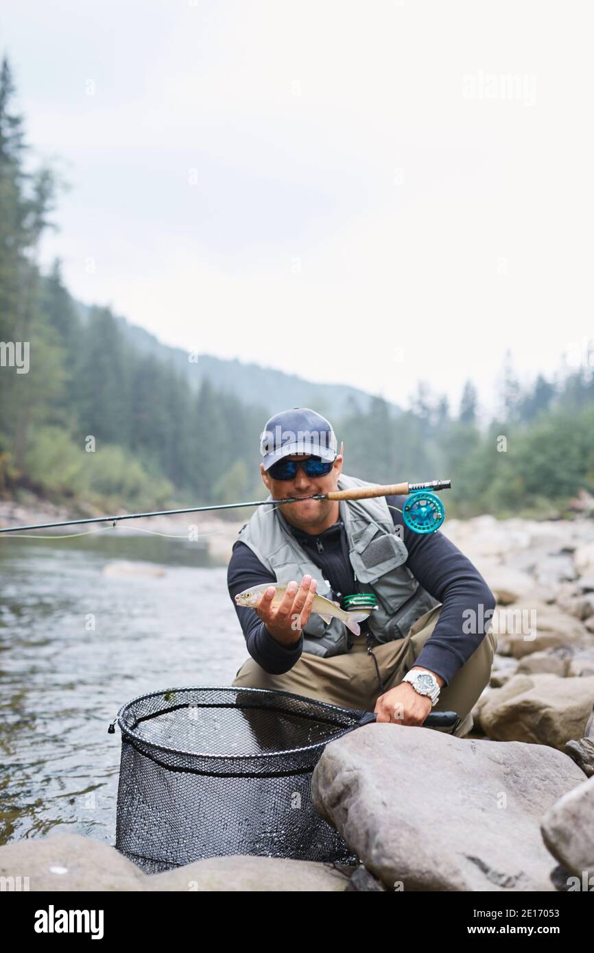 Smiling fisherman in casual clothing holding his rod in mouth and trout in  hands. Happy man sitting on stones among mountain river Stock Photo - Alamy