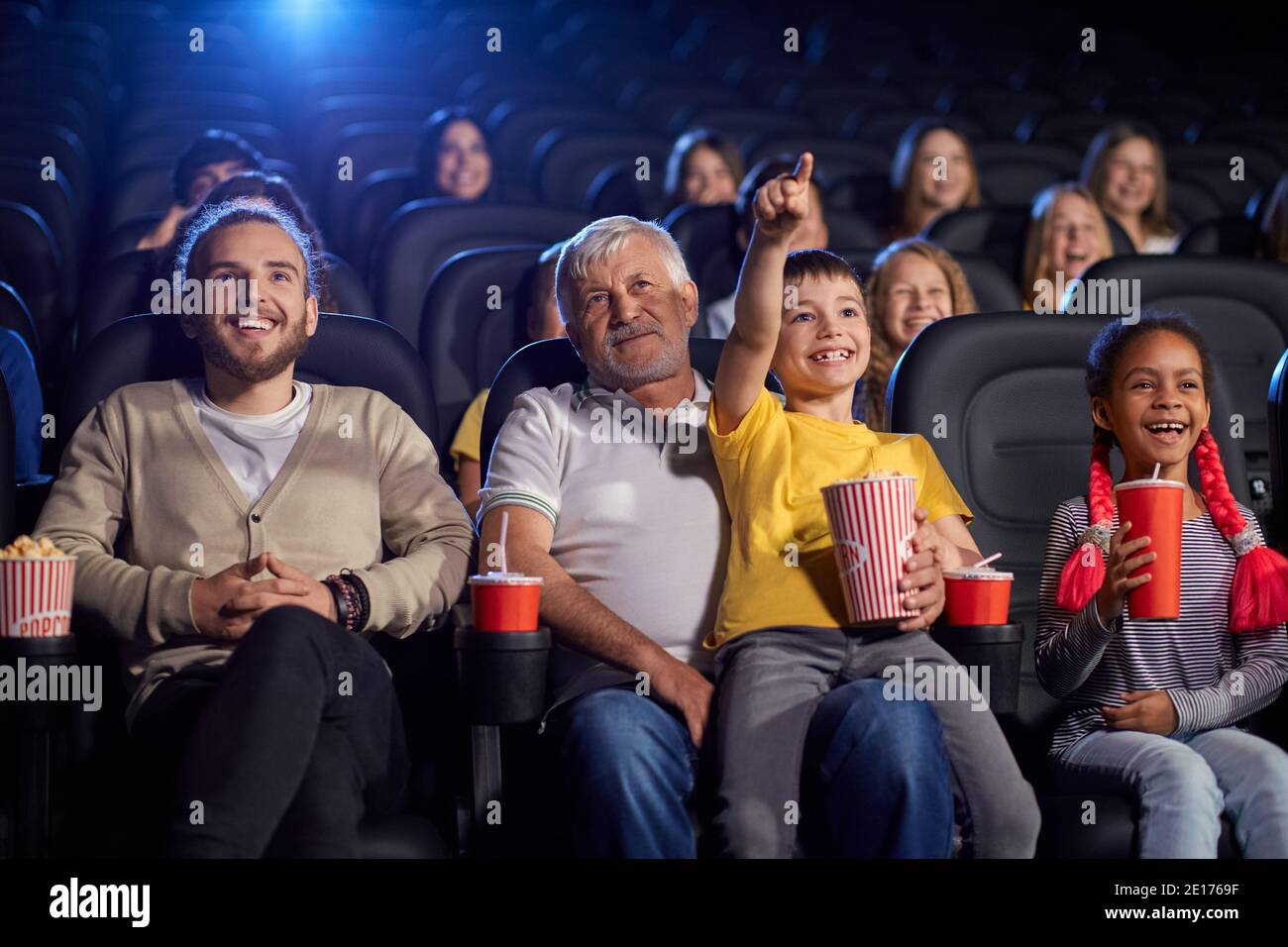 Bearded grandfather with grandson sitting on knees, child holding popcorn and pointing at screen, young multiracial audience laughing. Happy family enjoying time in cinema, watching funny cartoon. Stock Photo