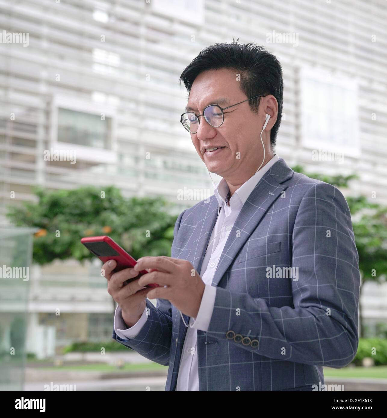 Asian businessman doing video call on smartphone using earphones in the street, smiling and looking at screen. Stock Photo