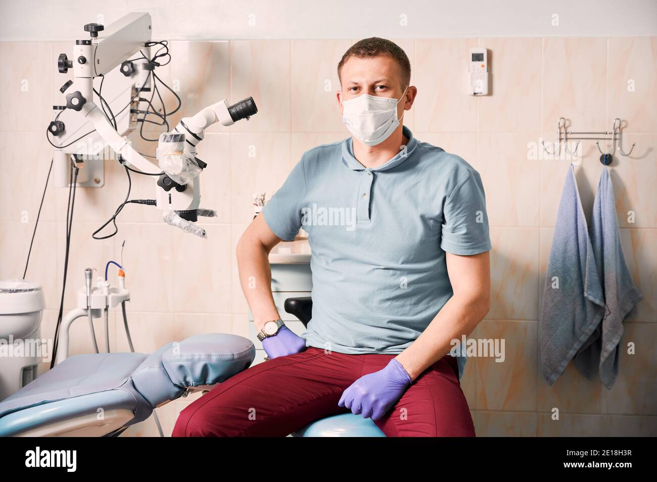 Portrait of man dentist in sterile gloves and medical mask looking at camera. Male stomatologist sitting near dental microscope. Concept of dentistry and stomatology. Stock Photo