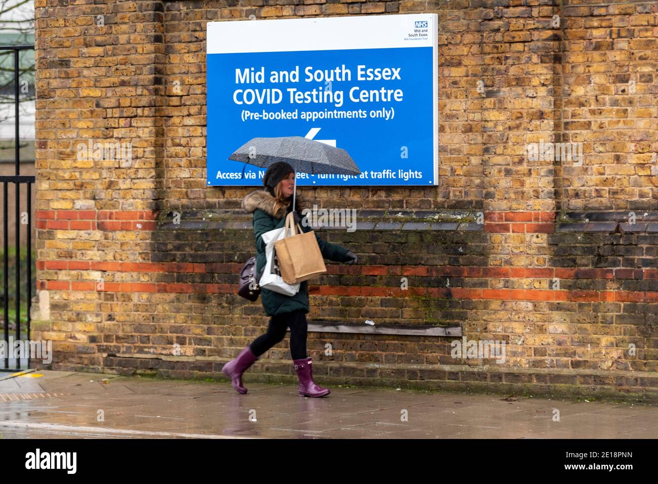 Southend on Sea, Essex, UK. 5th Jan, 2021. The seafront town of Southend on Sea has woken to a higher level of COVID 19 restrictions after Prime Minister Boris Johnson moved England into stricter tier 5 lockdown restrictions. People are out walking. A female with umbrella passing a sign for Mid and South Essex NHS COVID testing centre in heavy overcast wet weather Stock Photo