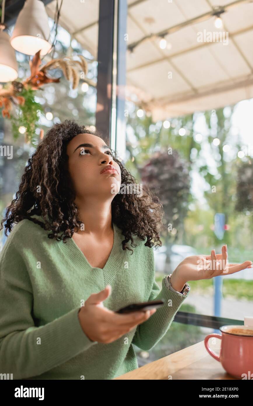 dissatisfied african american woman puffing cheeks, gesturing and holding smartphone while looking up in cafe Stock Photo