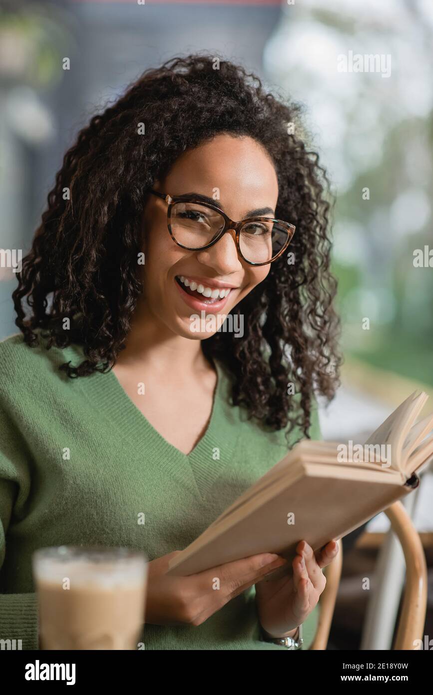 happy african american woman holding book while smiling in cafe Stock ...