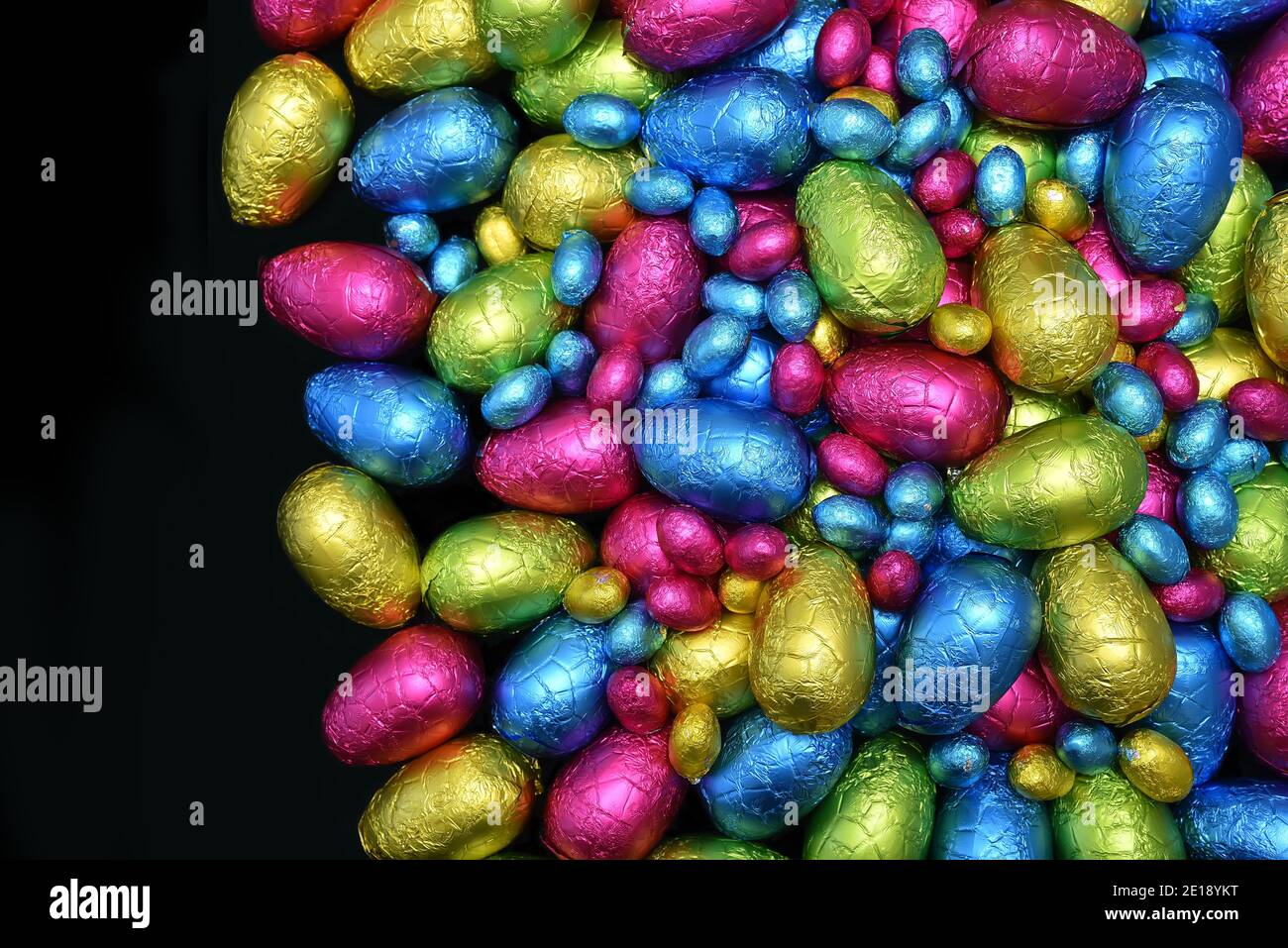 Pile or group of multi coloured & different sizes of colourful foil wrapped chocolate easter eggs in pink, blue, yellow and lime green against black. Stock Photo