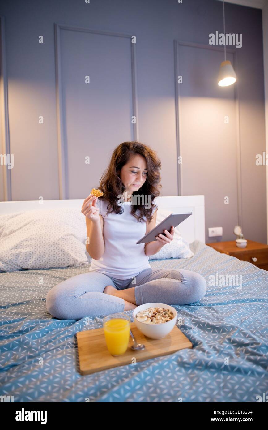 Pretty satisfied middle aged women sitting on the bed and looking on a table before sleeping while eating a biscuit. Stock Photo