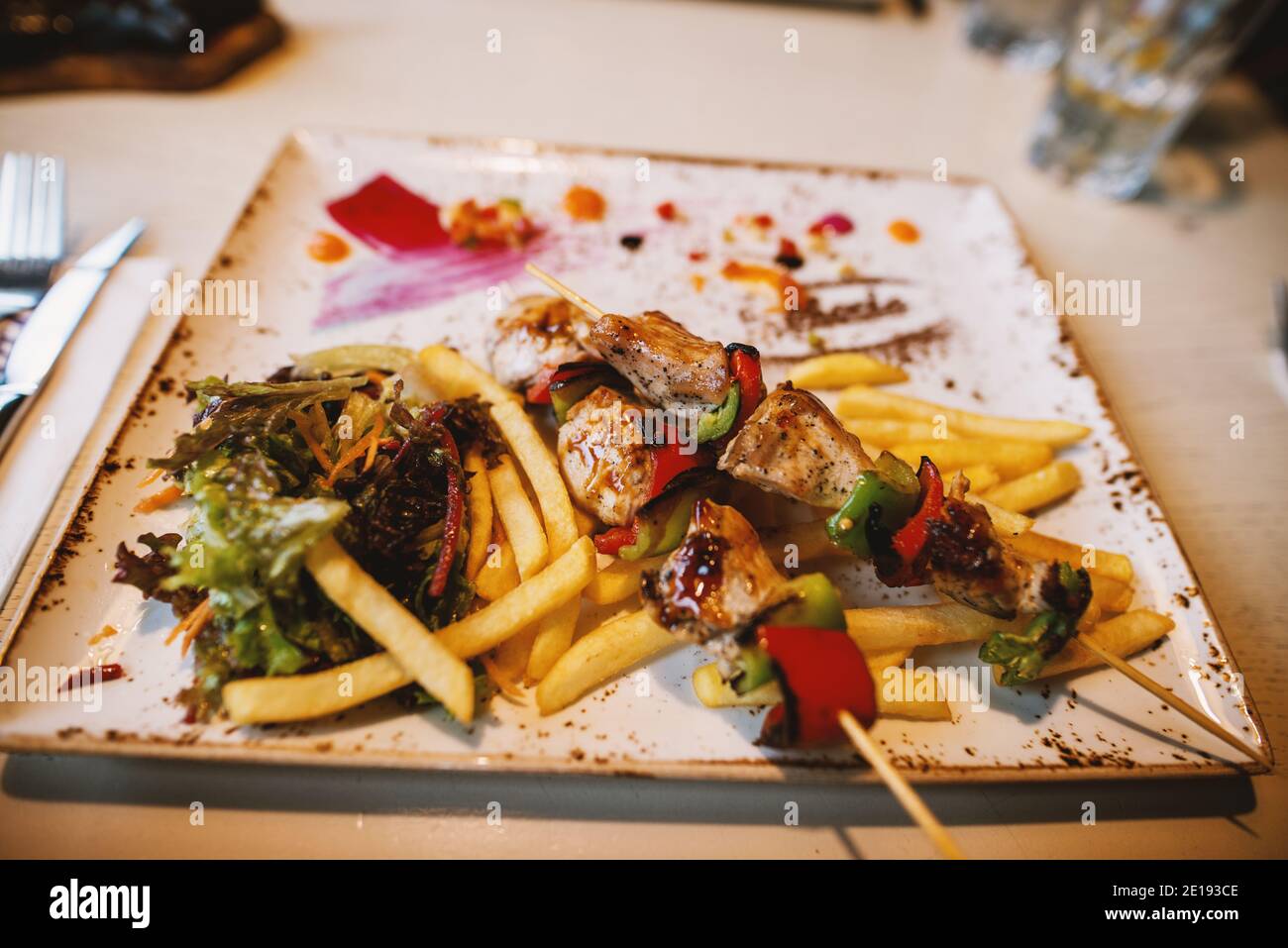Close up focus view of fried skewers on a wooden stick and chips with salad on the plate in the restaurant. Stock Photo