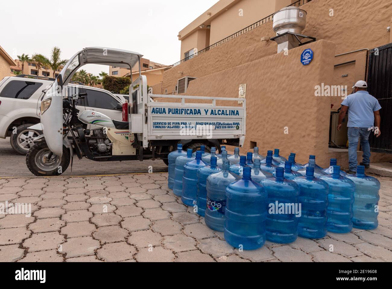 A man dressed in blue jeans and blue shirt delivers bottles of purified water in his trike cargo truck, large blue bottles lined up on the pavement. Stock Photo