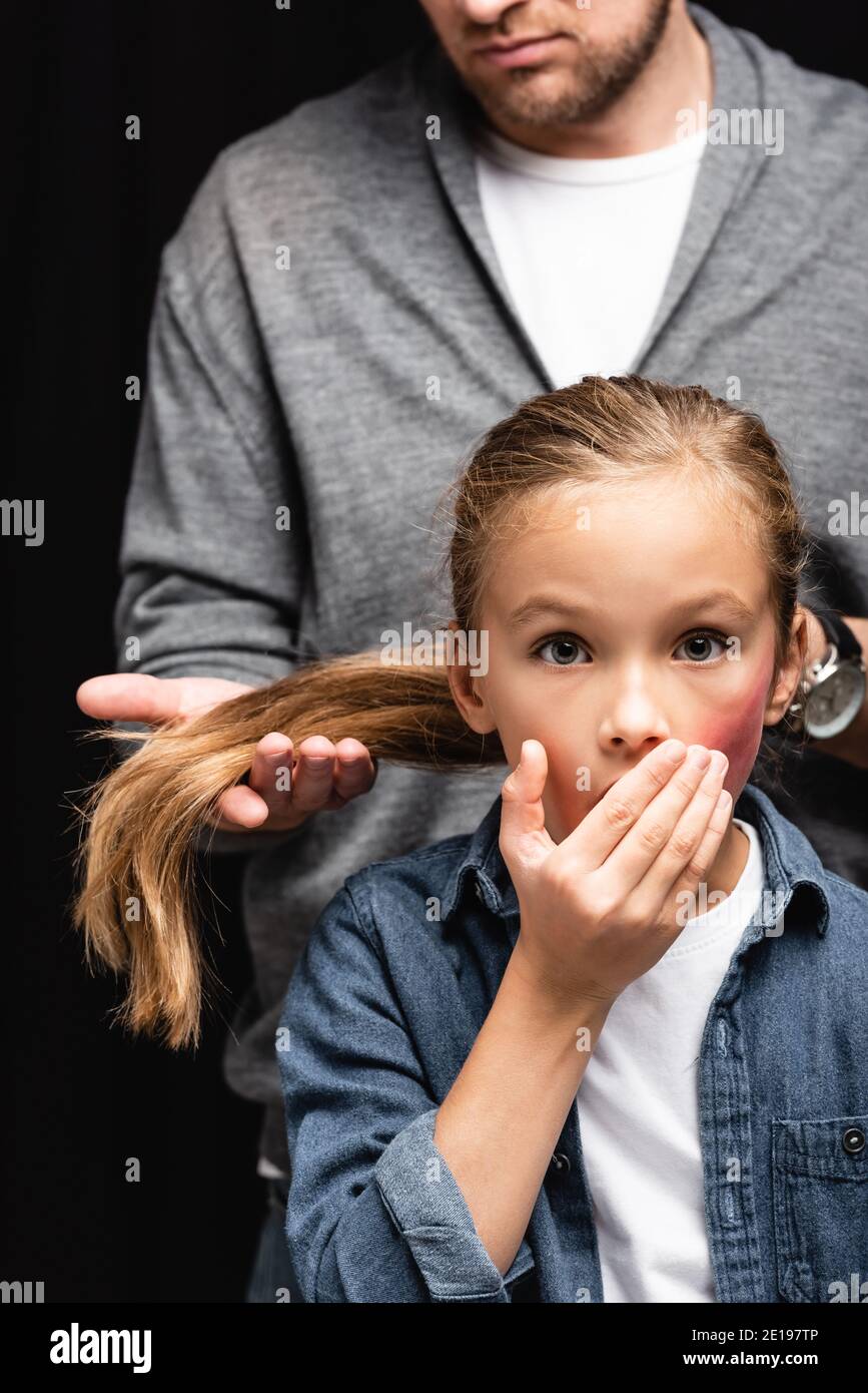 Scared kid with bruise on face standing near abusive father touching hair on blurred background isolated on black Stock Photo