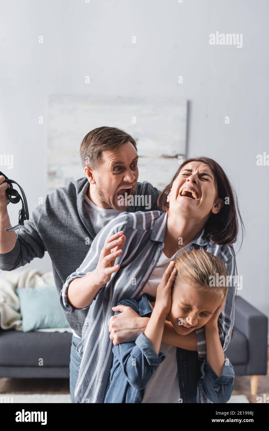 Crazy man holding waist belt near crying wife and child at home Stock Photo