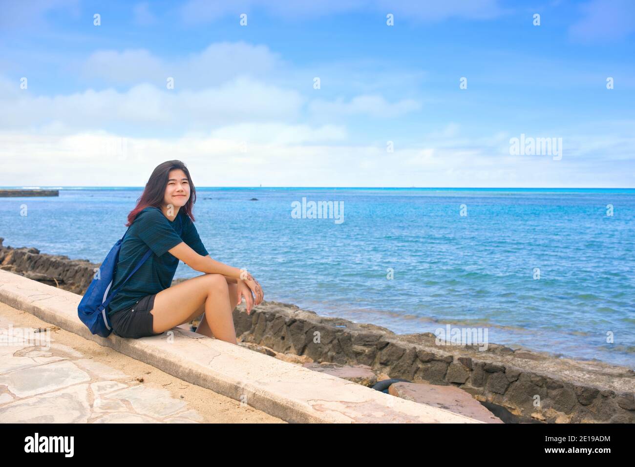 Biracial teen girl sitting on concrete walkway next to blue ocean in tropical Hawaii with blue skies and horizon in background Stock Photo