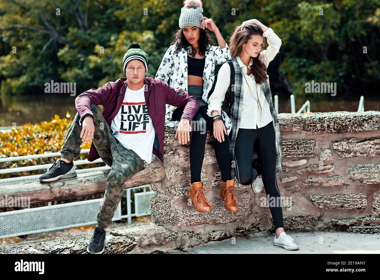 Portrait of 3 young adults sitting on a tree log in front of a lake at a natural park during a sunny day in the spring wearing cool urban outfits. Stock Photo