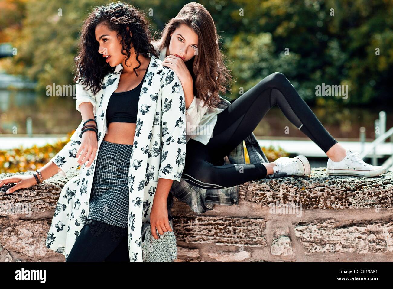 Portrait of 2 young adult women lounging on a stone wall in front of a lake at a natural park during a sunny day wearing cool urban outfits. Stock Photo