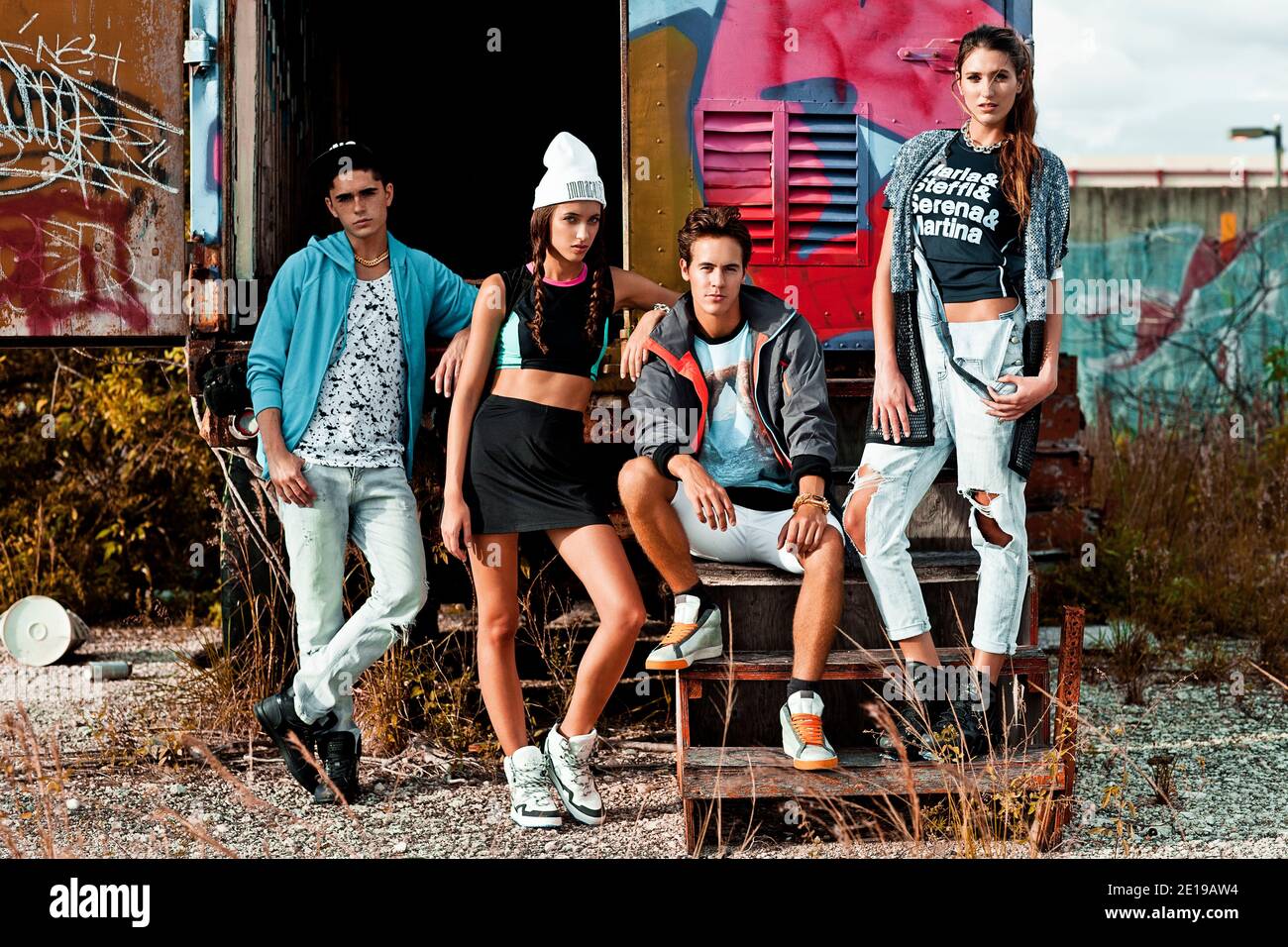 A portrait of an interracial group of 4 young adults lounging on wooden steps in front of an open graffiti sprayed container on a vacant lot. Stock Photo