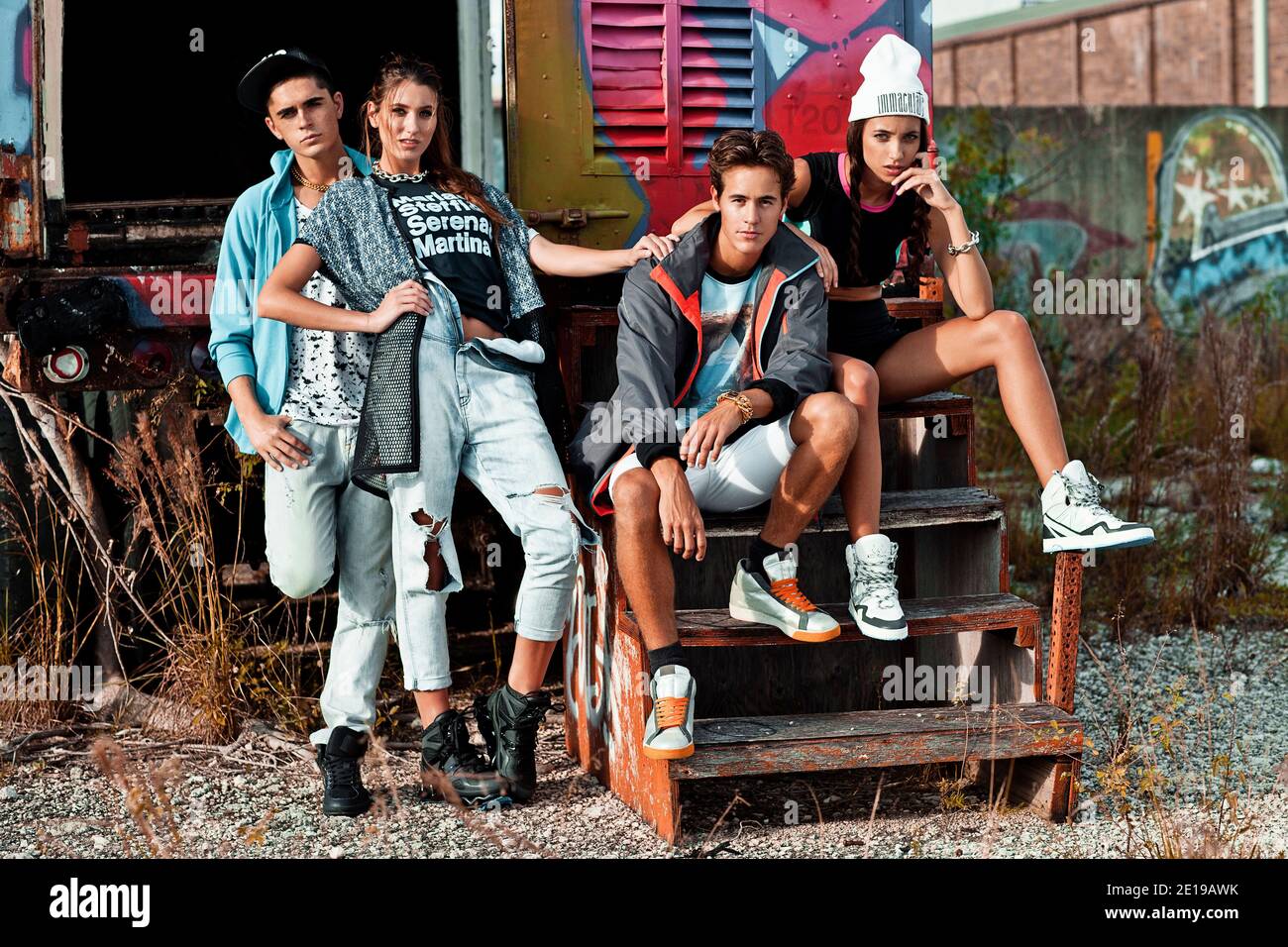 A portrait of an interracial group of 4 young adults sitting on wooden steps in front of an open graffiti sprayed container on a vacant lot. Stock Photo