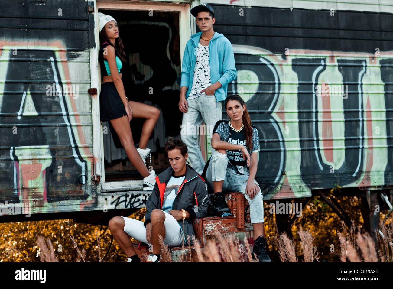 A portrait of an interracial group of 4 young adults lounging on wooden stairs in front of an open graffiti sprayed container on a vacant lot. Stock Photo