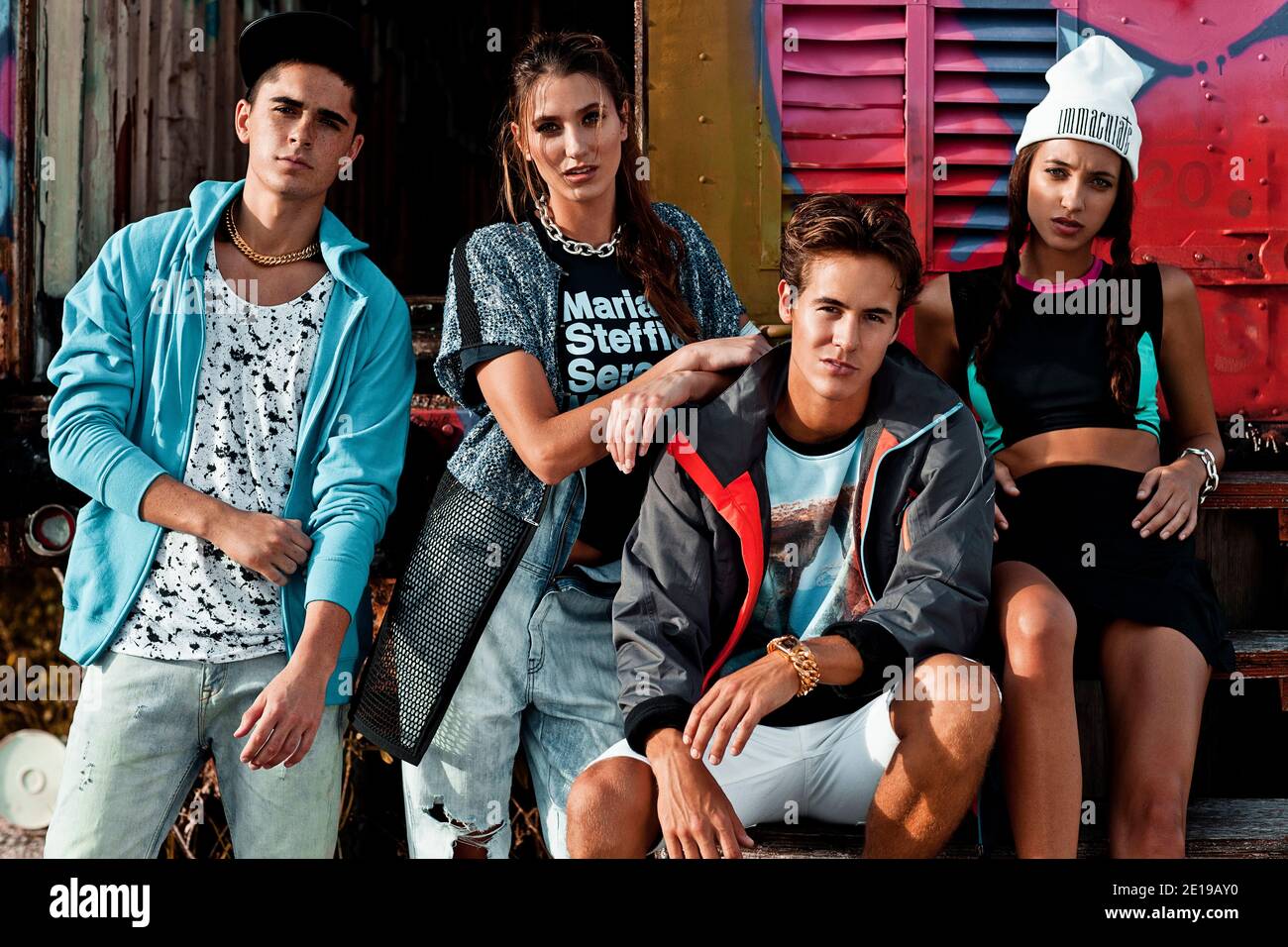 Closeup portrait of an interracial group of 4 young adults sitting on wooden steps in front of an open graffiti sprayed container on a vacant lot. Stock Photo