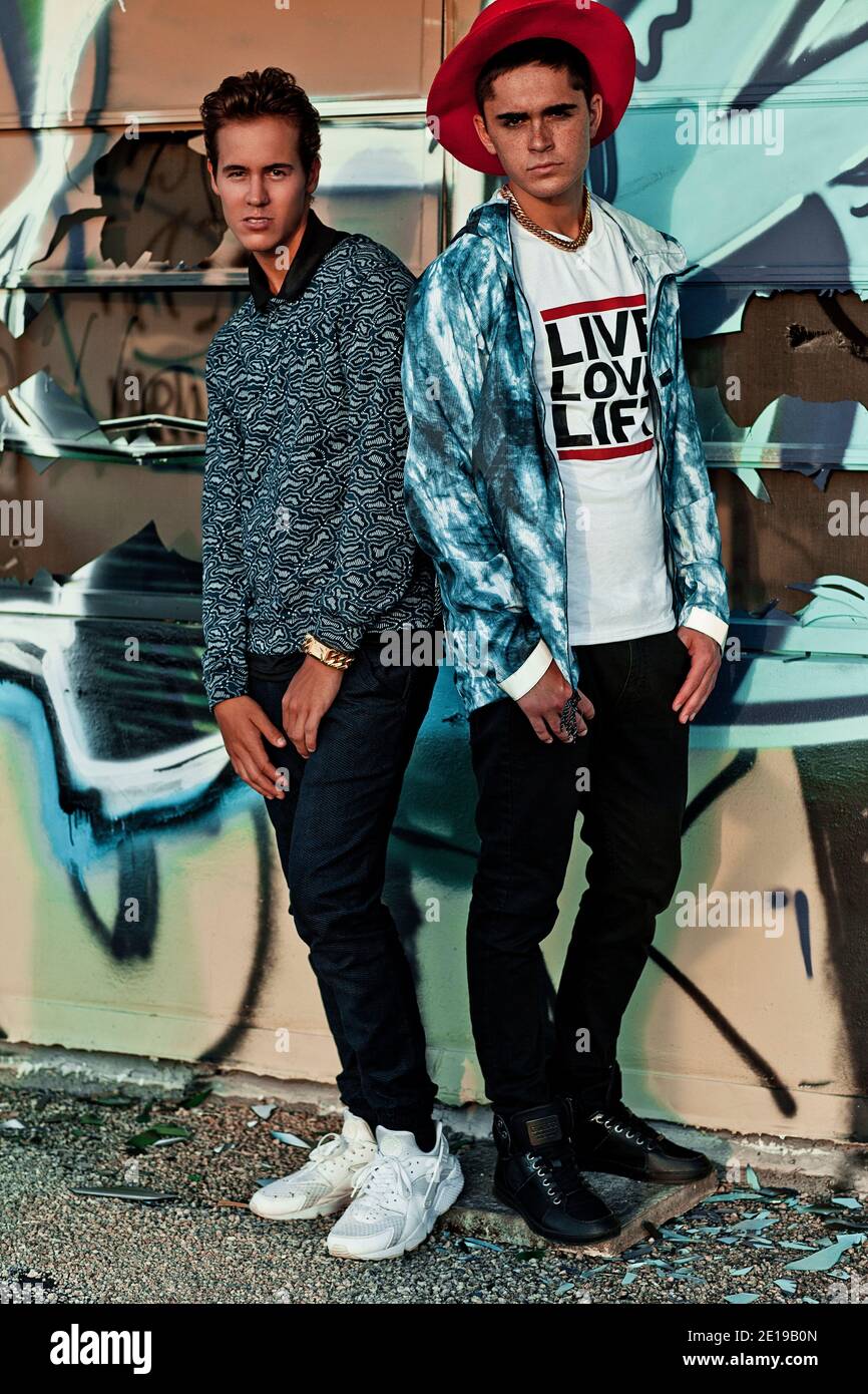 2 young male adults standing in front of a graffiti sprayed building with broken windows on a vacant lot on a warm summer day wearing urban outfits. Stock Photo