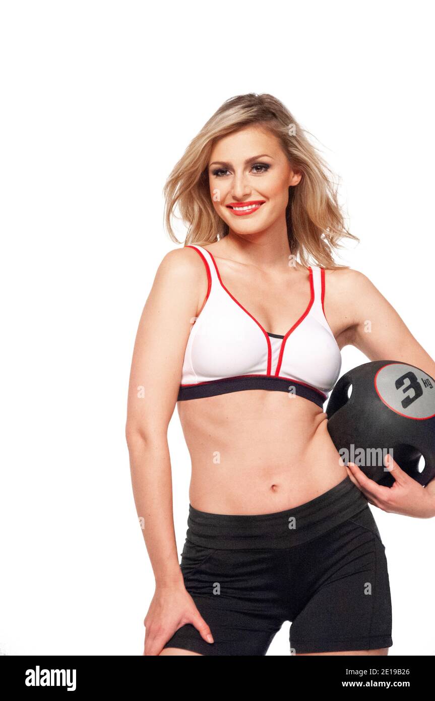Portrait of a fit white female athlete with curly long blond hair posing by herself holding a black medicine ball in a studio with white background. Stock Photo