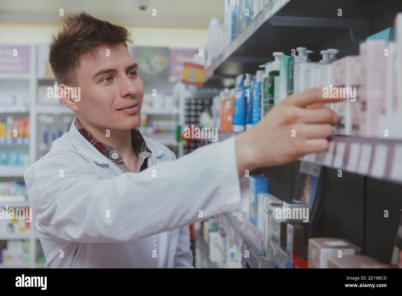 Cheerful handsome male pharmacist doing stocktaking, organizing products on shelves at drugstore. Charming young male chemist working at the pharmacy. Stock Photo