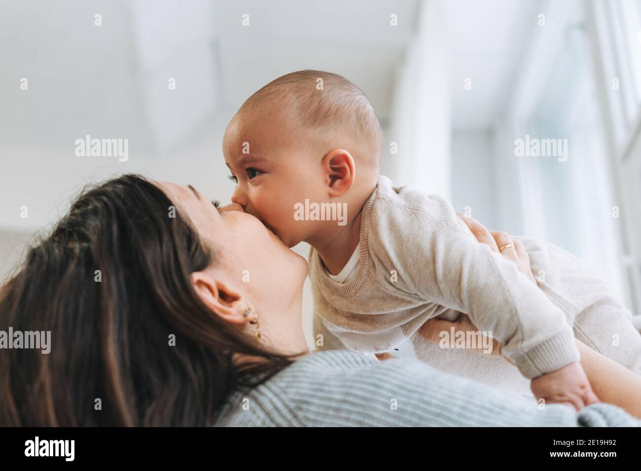 Young mother kissing her cute baby boy on hands in bright room ...