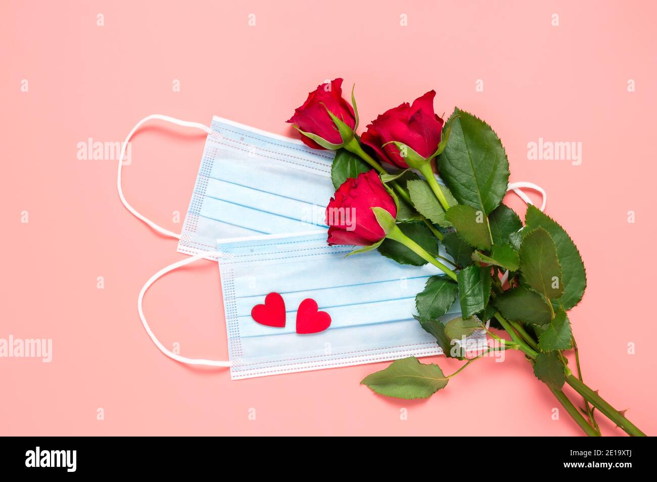 Red roses and disposable face masks with two hearts on pink background. Mothers, womens or Valentines day celebration during coronavirus pandemic Stock Photo
