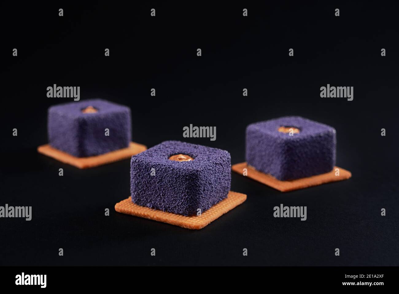 Closeup view of matte purple pie pieces filled with brown creme isolated on black studio background. Three small square matte cakes in row on cookies in restaurant. Sweet bakey, desserts concept. Stock Photo