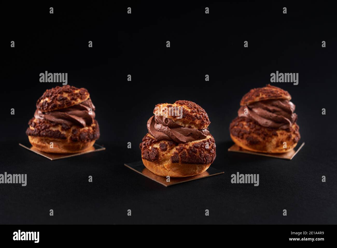 Three delicious fresh crunchy profiteroles with sweet brown chocolate cream inside. Closeup of homemade tasty baked eclairs isolated on black background. Concept of desserts, restaurant food. Stock Photo