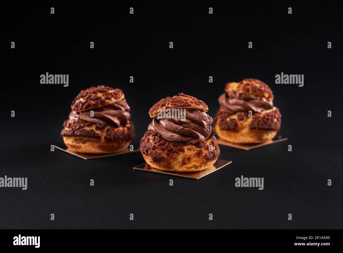 Three delicious fresh crunchy profiteroles with sweet brown chocolate creamy filling. Closeup of homemade tasty baked eclairs isolated on black background. Concept of desserts, restaurant food. Stock Photo