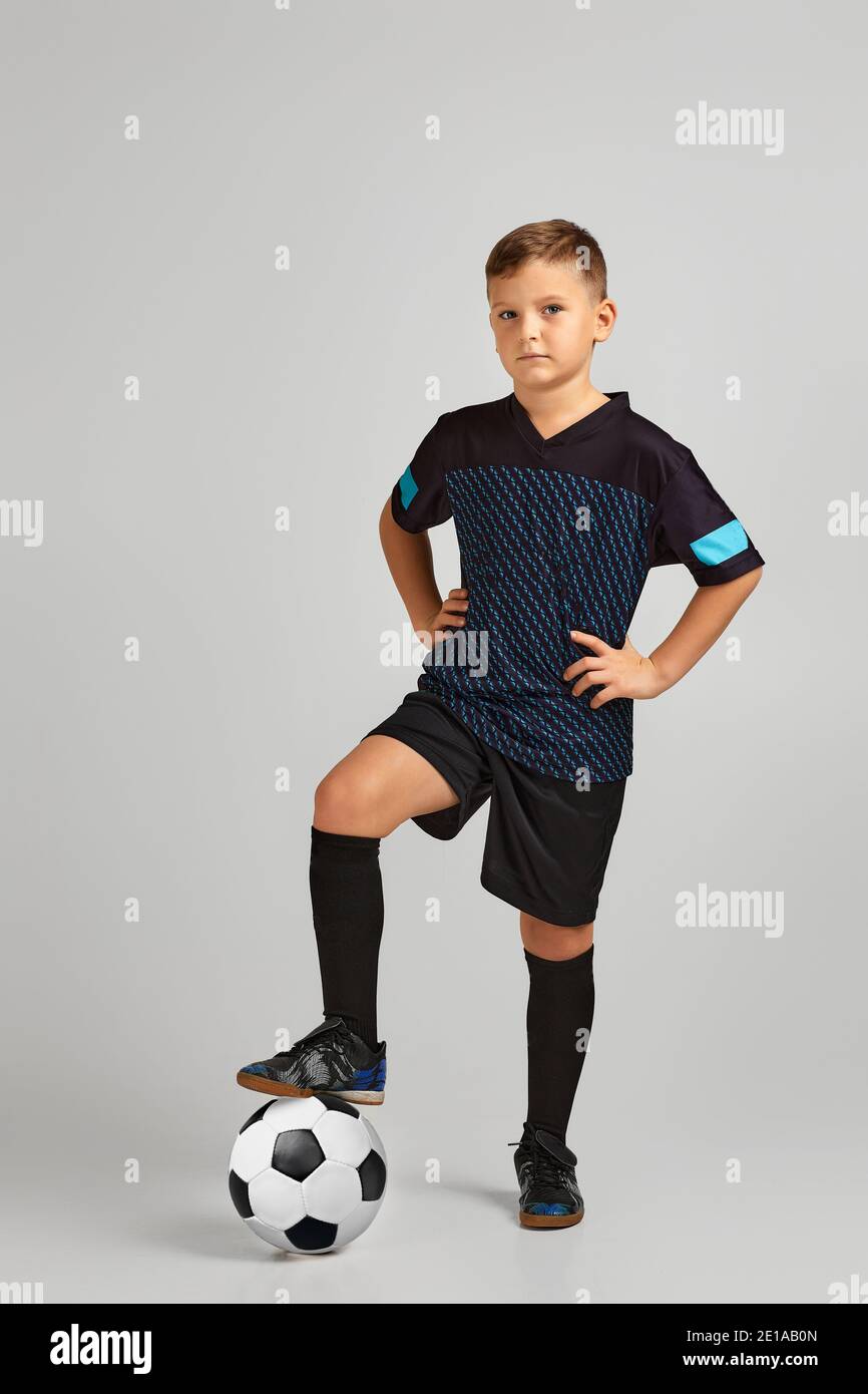 little boy football player in uniform holding his leg on ball over studio background. child dreams of becoming a soccer player. Stock Photo