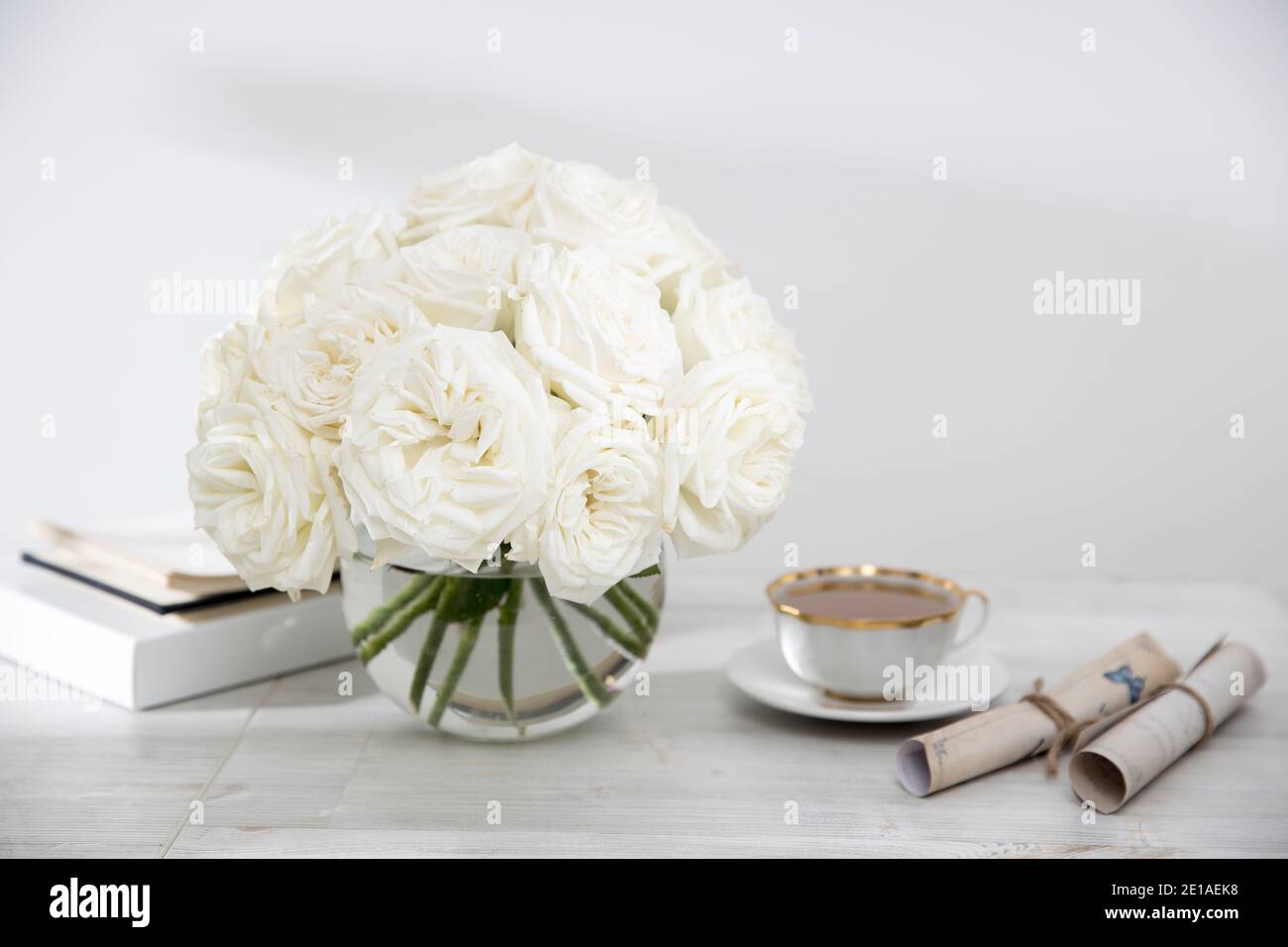 A bouquet of white roses in a round glass vase on a table with cup of ...