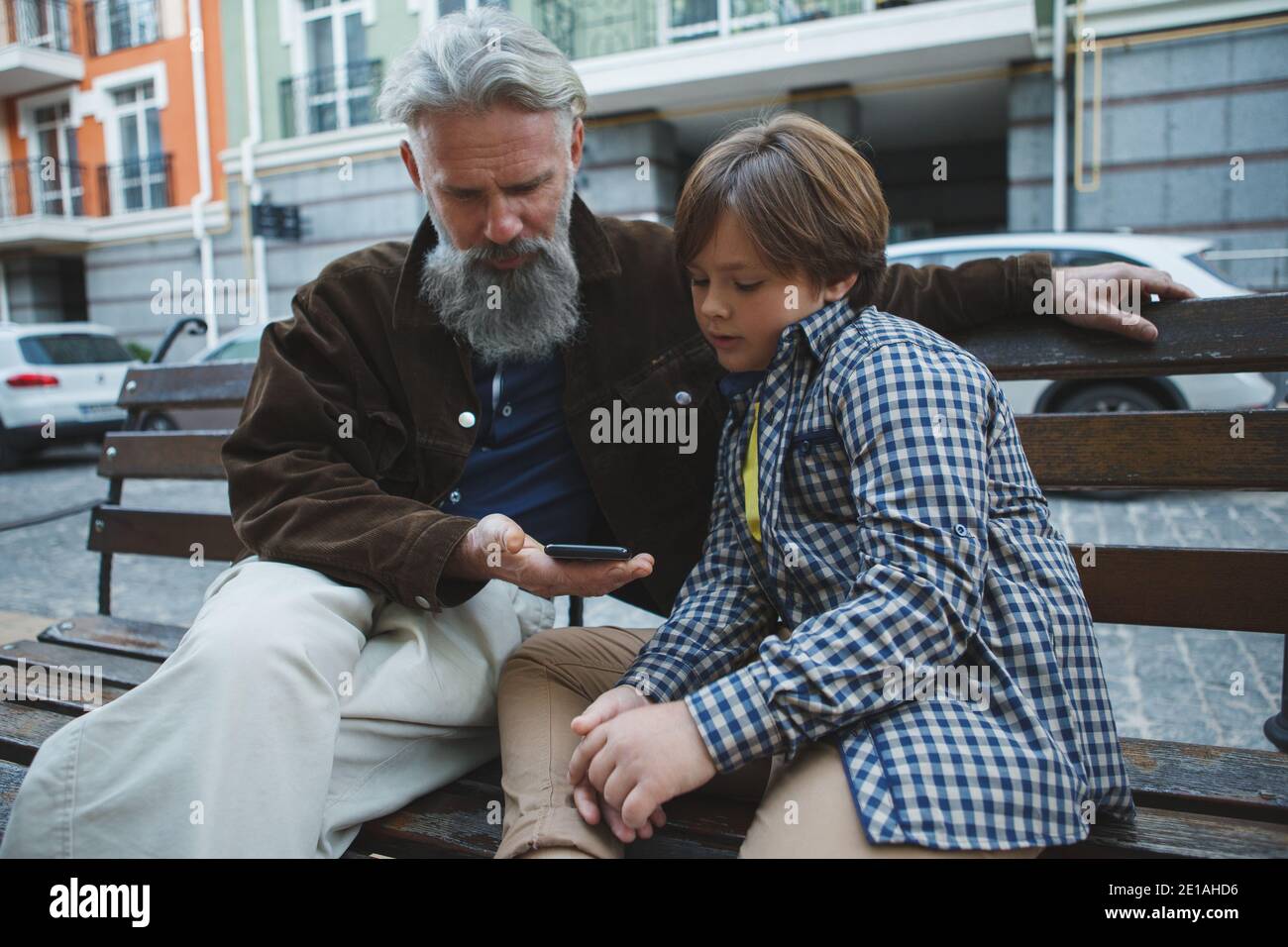 Elderly man using smart phone with the help of his young grandson, sitting on a bench in the city Stock Photo