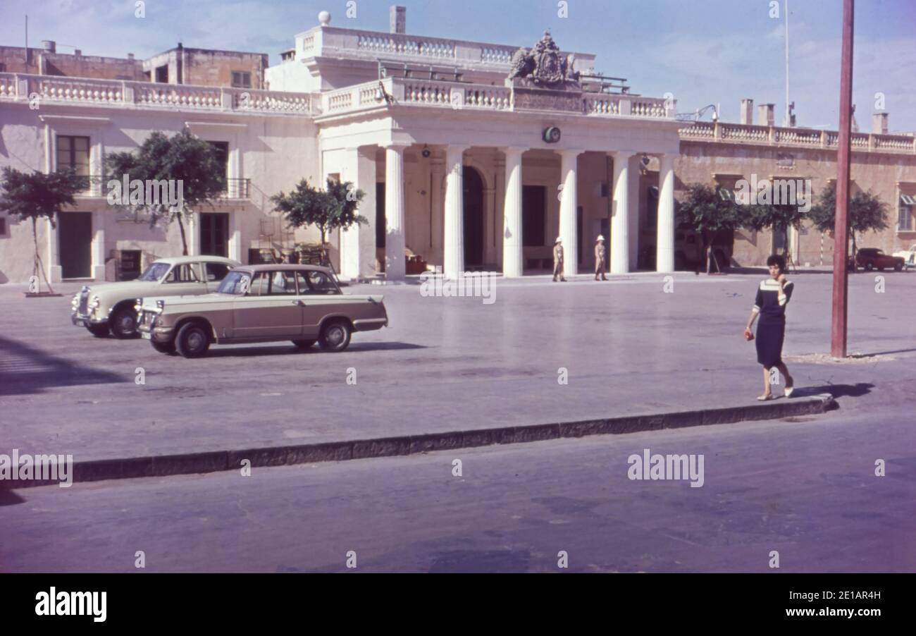 A stylish dressed woman walking in front of the Main Guard on St George’s Square Valletta, Malta. Triumph Herald and MG Magnette cars are parked. 1959 1960. Scanned from slide. Stock Photo