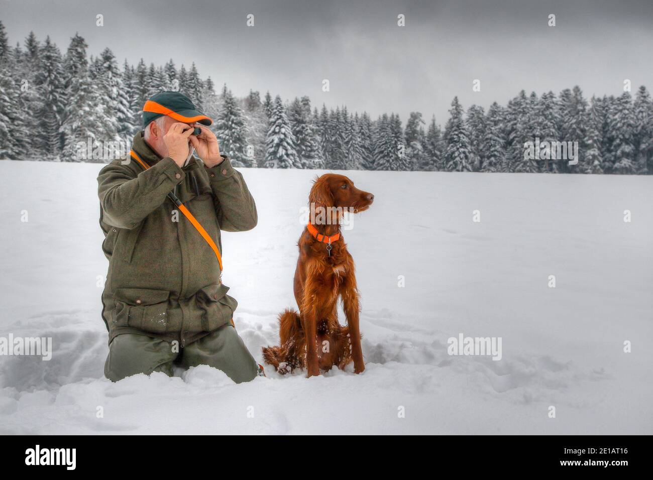 In winter a hunter kneels in the snow and observes his snowy hunting ground through a small eyepiece, next to him sits his Irish Setter dog. Stock Photo