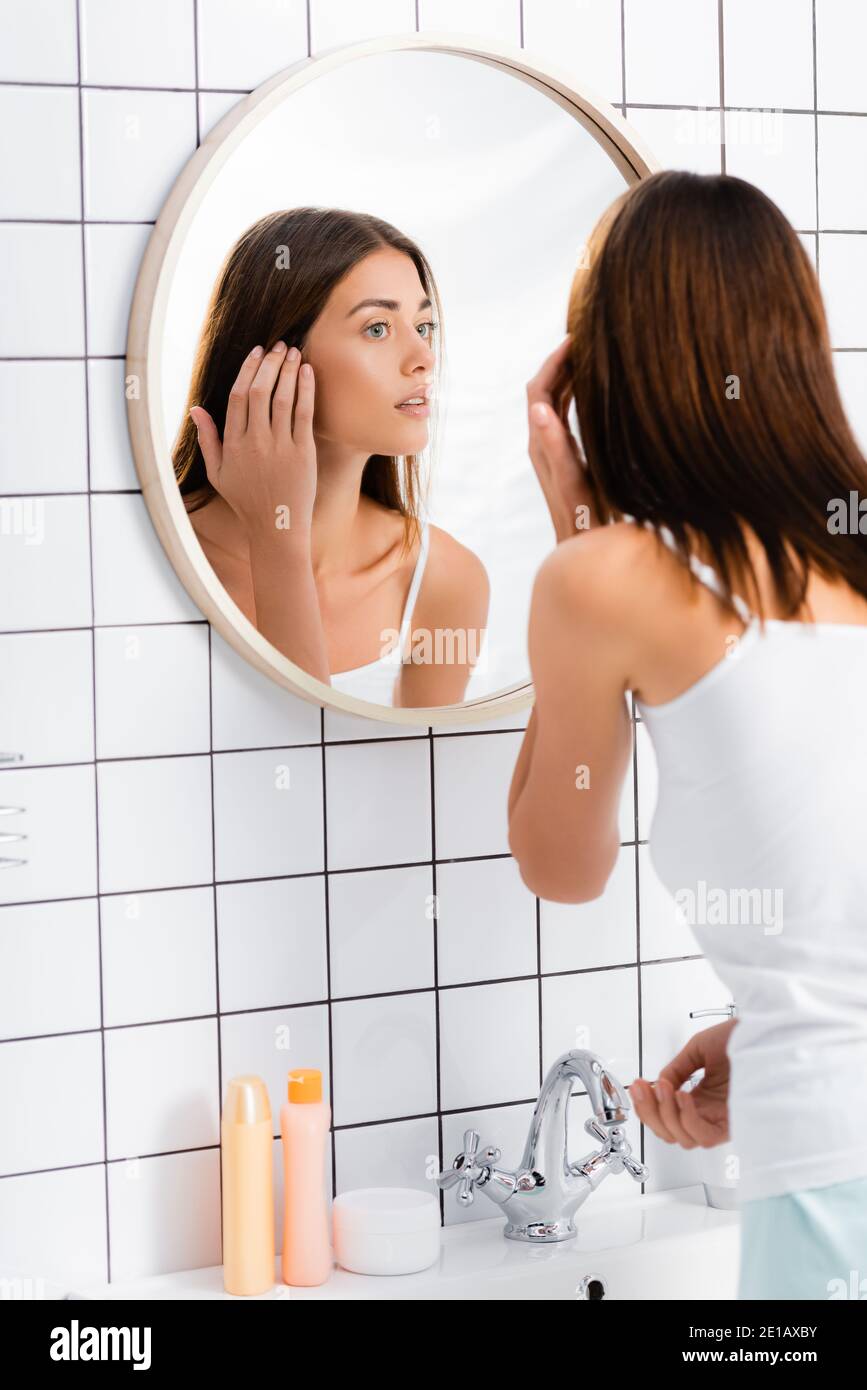 young woman in white singlet looking in mirror in bathroom Stock Photo