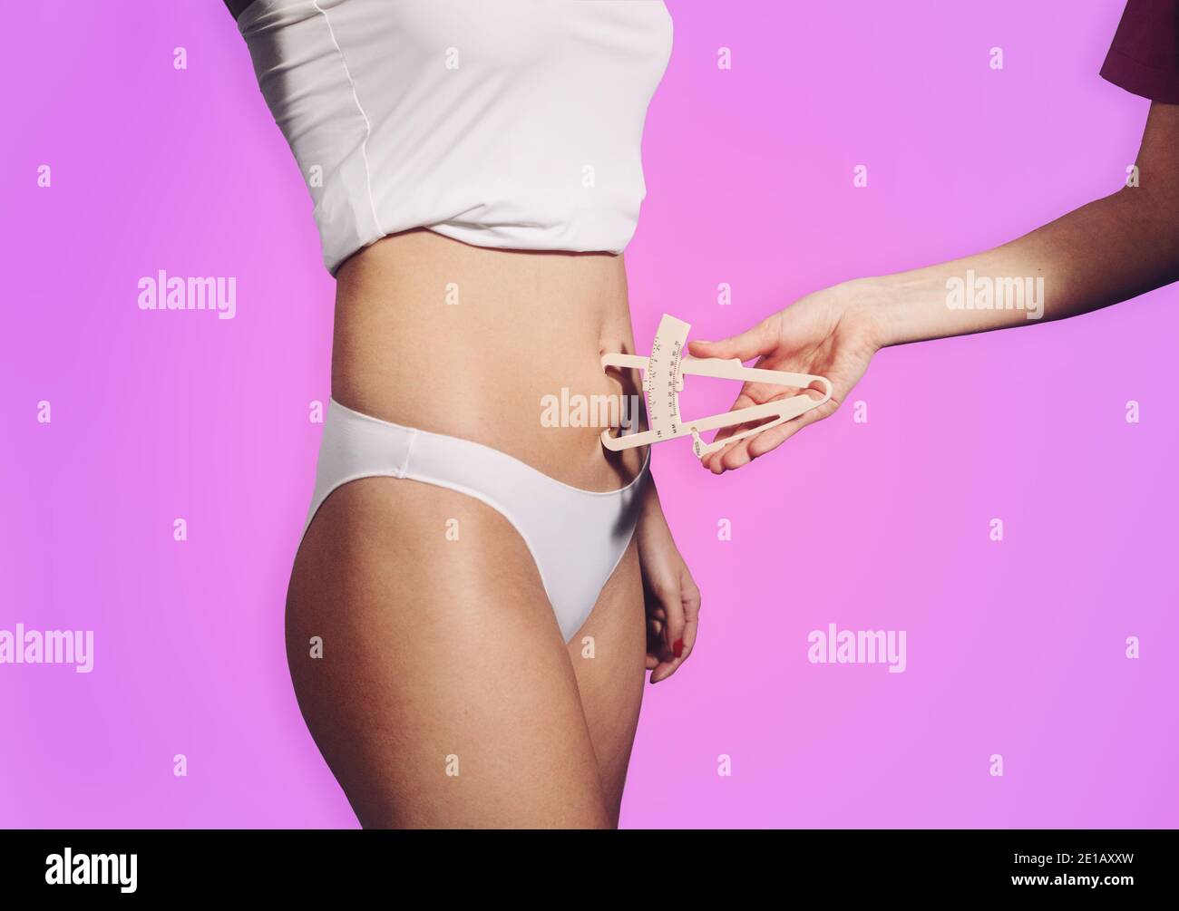 Closeup shot of young attractive woman's belly. Nutritionist hand measuring body fat waist layer with caliper on pink gradient background Stock Photo