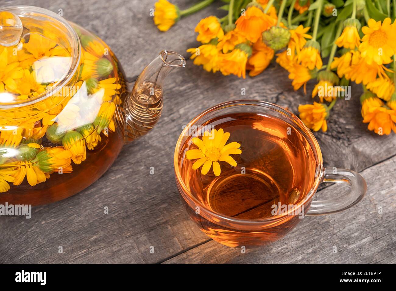 Cup of herbal tea and transparent teapot and marigold flowers on wood