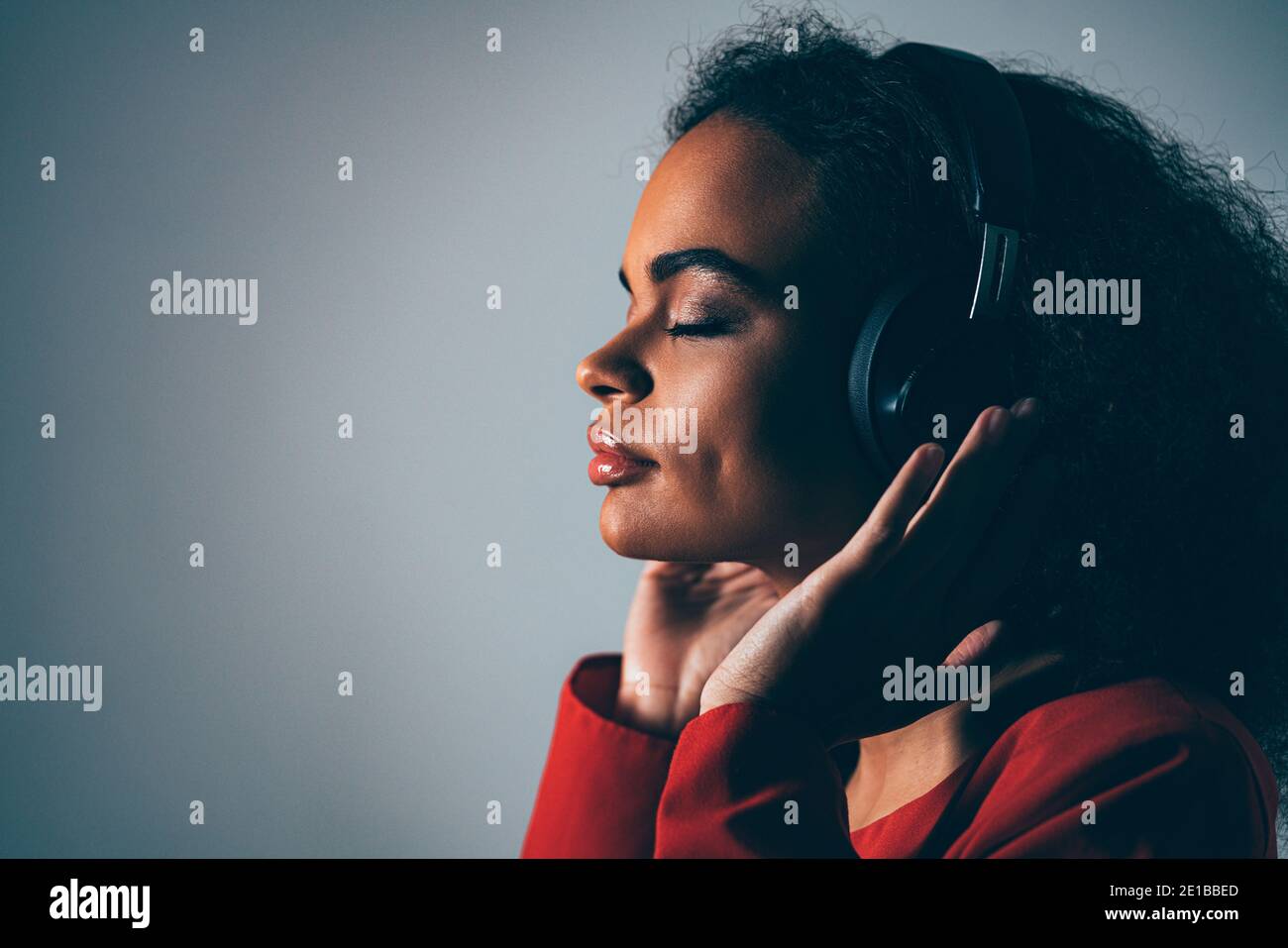 Joy of a music young African-American girl listening her favourite track in headphones wearing red jacket black top under isolated on grey background Stock Photo