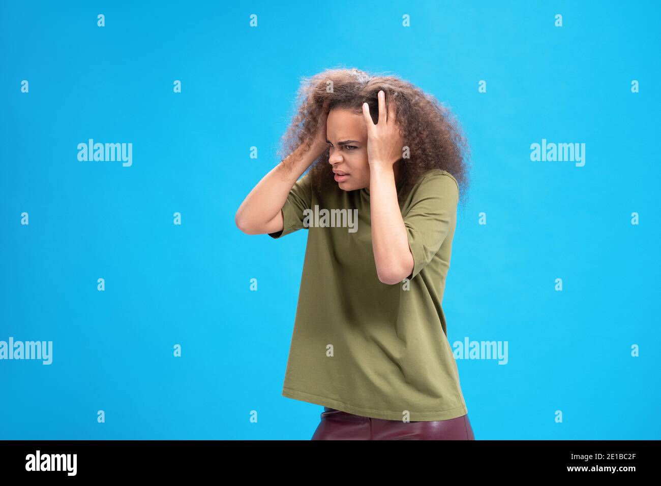 Experiencing migraine or headache African American young woman standing holding head with hands in olive t shirt isolated on blue background. Human Stock Photo