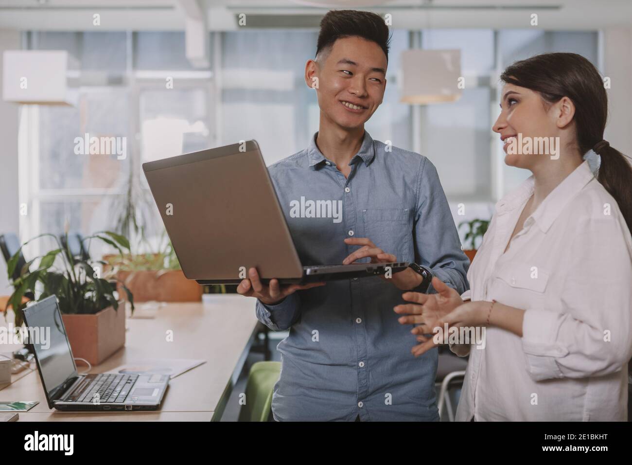 Young businesspeople laughing, talking at work, using laptop tigether. Asian businessman discussing ideas with his female colleague, copy space. Inter Stock Photo