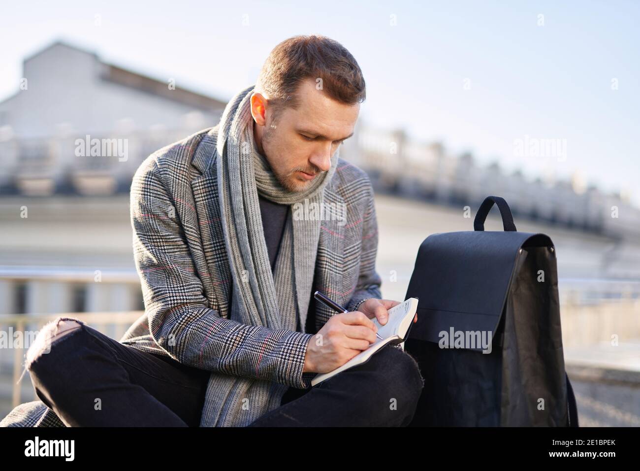 Caucasian bearded man in scarf and grey coat making notes outdoor. Male thinking and working on a sunny spring or autumn day. Working and studying con Stock Photo