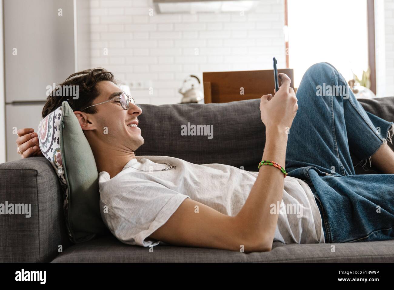 Picture of optimistic pleased handsome man lies on sofa indoors at home using mobile phone Stock Photo