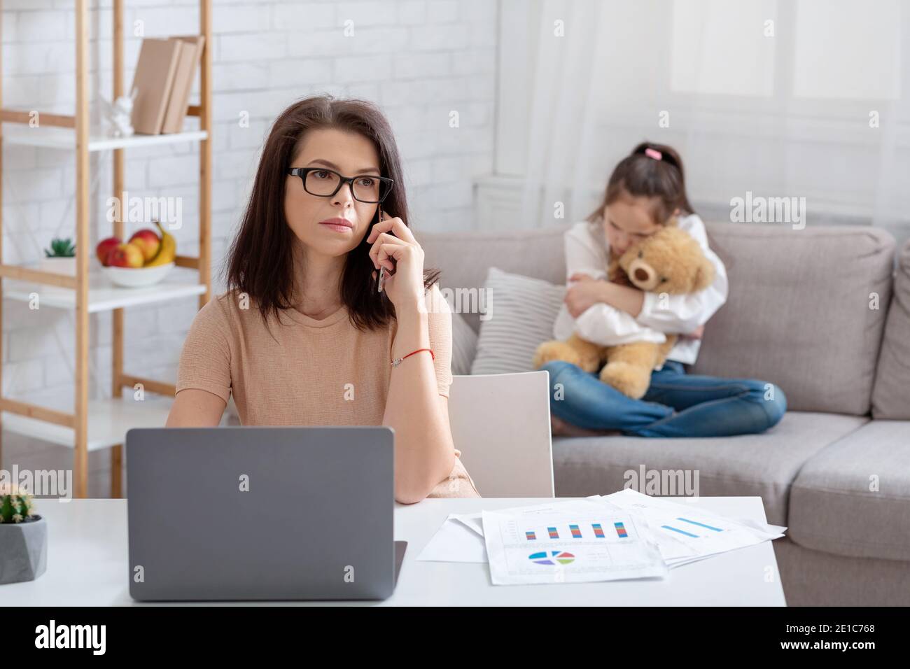 Modern working mom doing online job, her neglected daughter hugging teddy bear, feeling lonely and sad at home Stock Photo