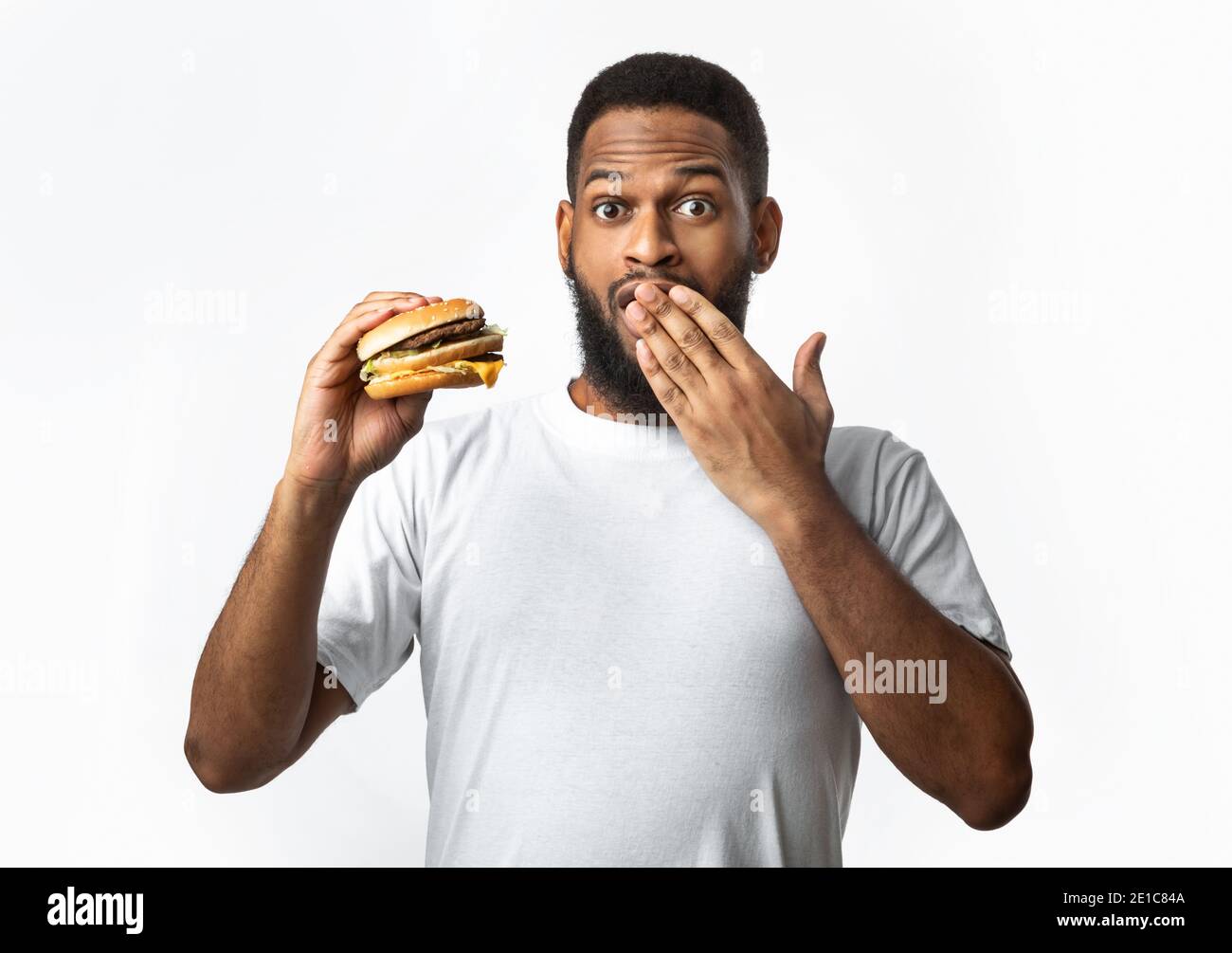 Funny African Guy Holding Burger Covering Mouth On White Background