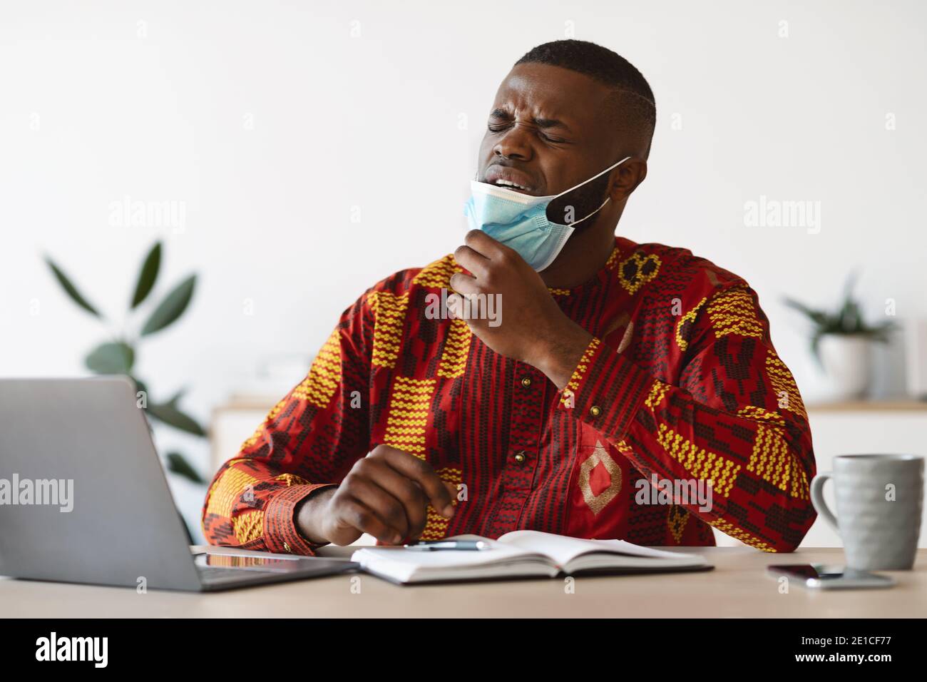 Young African Man Taking Off Medical Mask At Work, Can Not Breathe Stock Photo