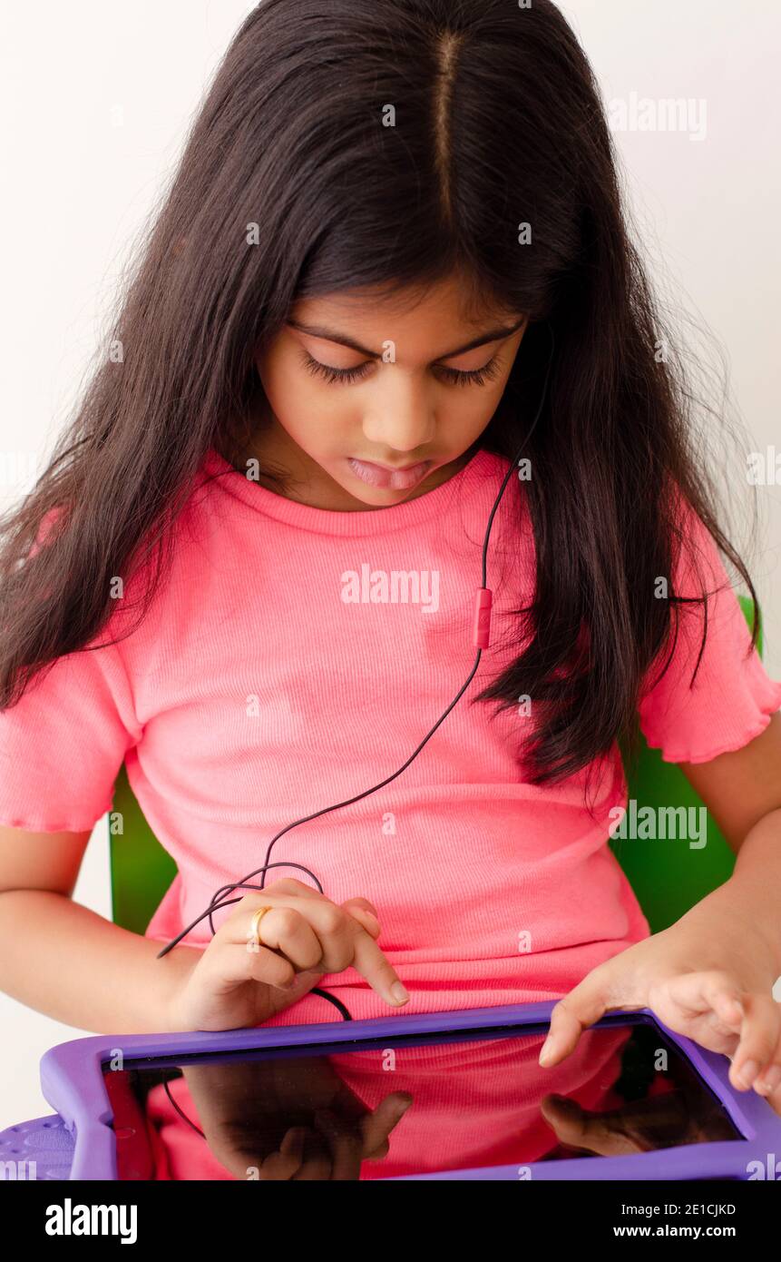 A little Indian girl  with earphones and tablet taking online classes during covid pandemic. e-learning concept. Stock Photo