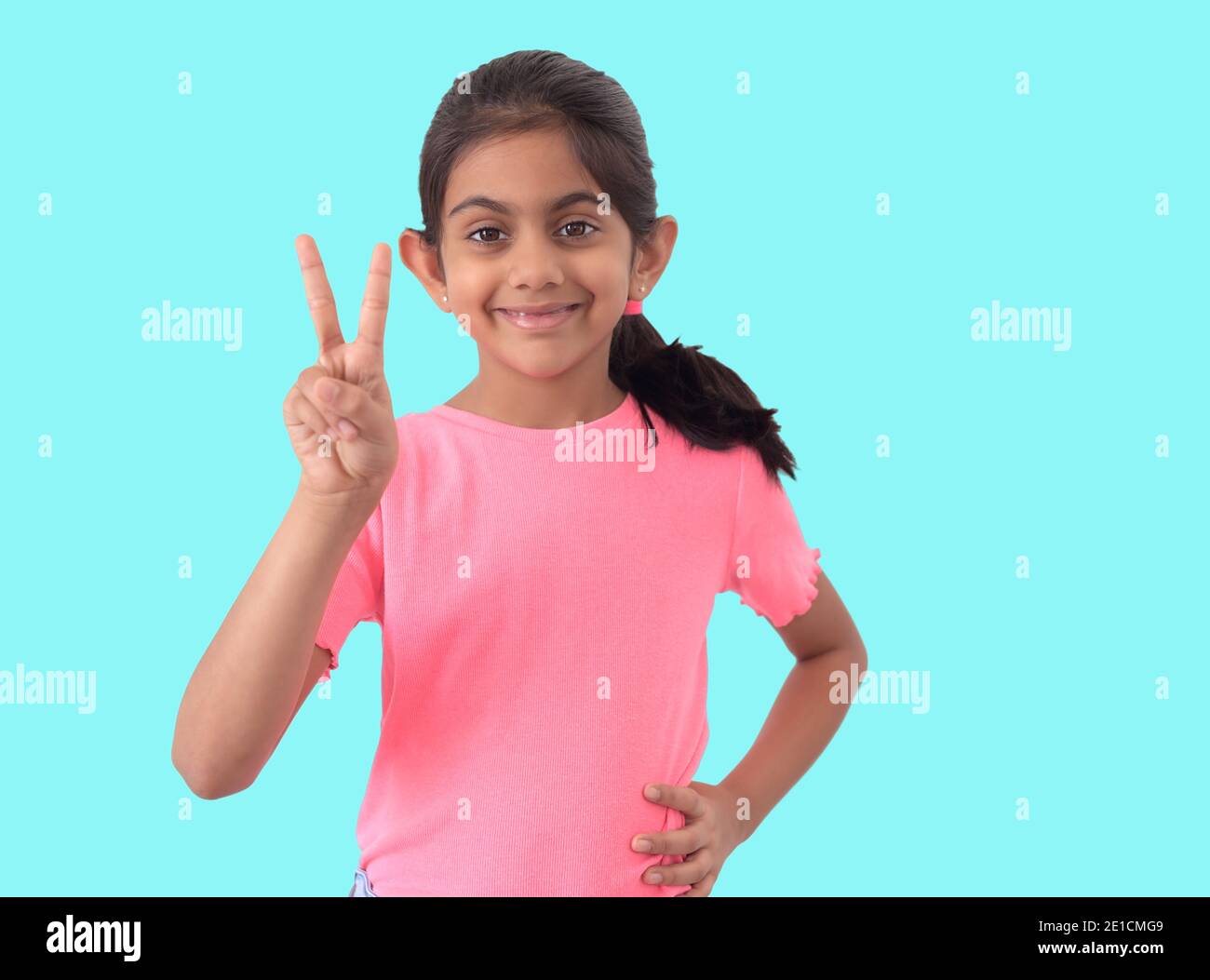 Portrait of a beautiful and smart Indian girl showing victory or winning symbol using hand in blue background while smiling ,confident and happy. Stock Photo