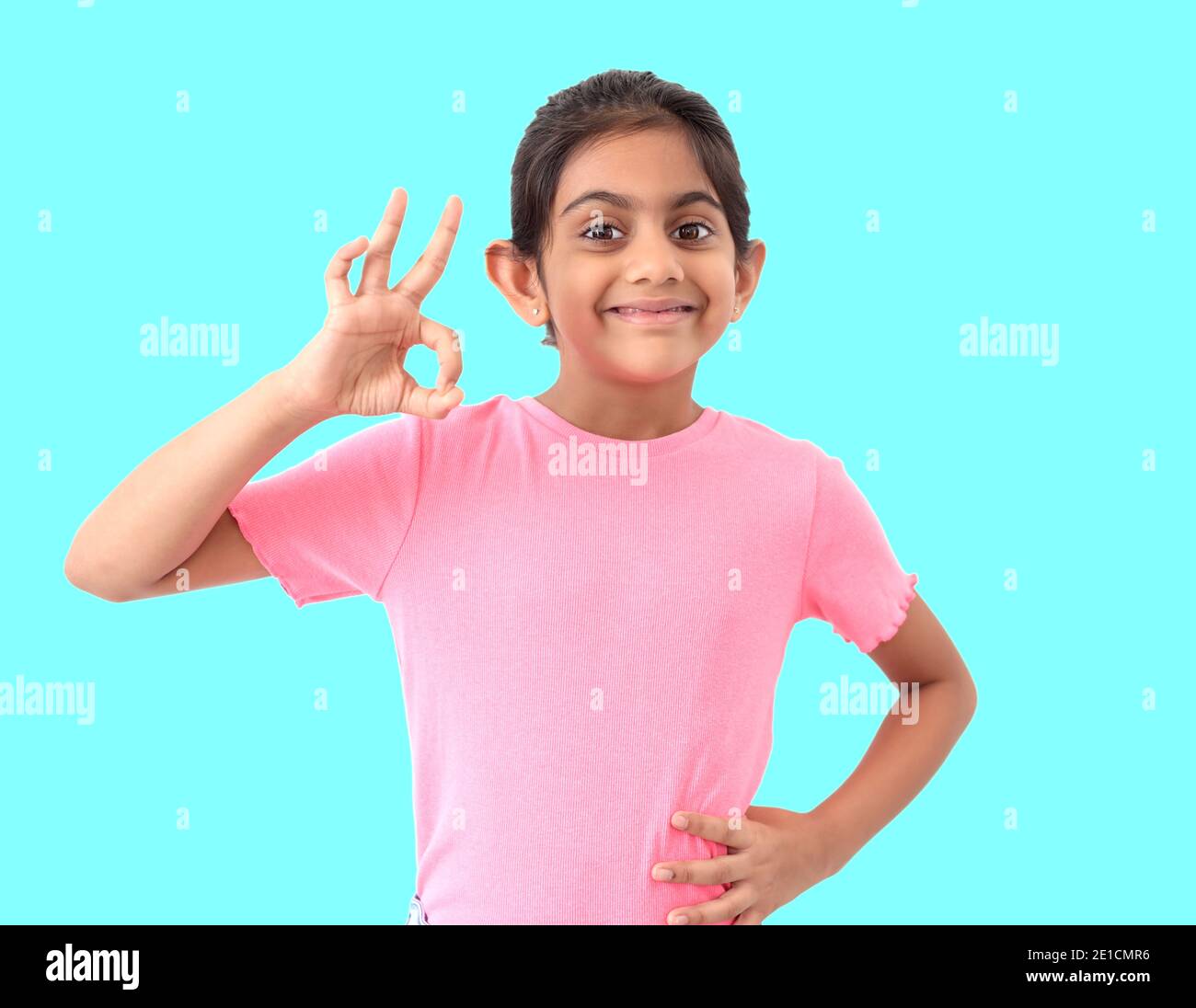 Portrait of a cute little indian girl in blue background showing 'perfect ' shape sign with hands and expressing satisfaction and appreciation. Stock Photo