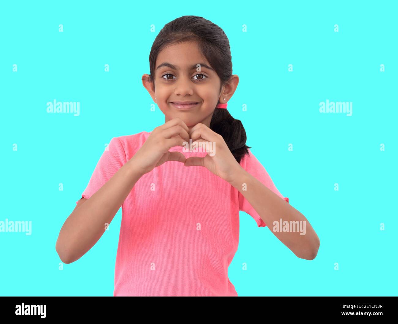 Portrait of a cute little indian girl showing heart shape sign with hands and expressing love in blue background. Stock Photo