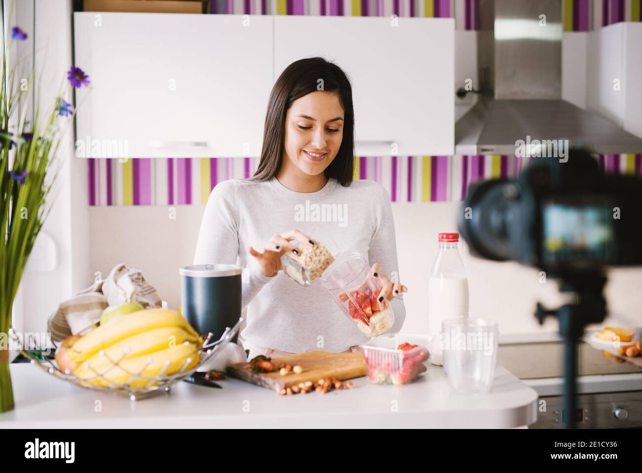 Young beautiful girl is putting cereals in a mixer bowl that is already filled with fresh fruit in front of the camera. Stock Photo